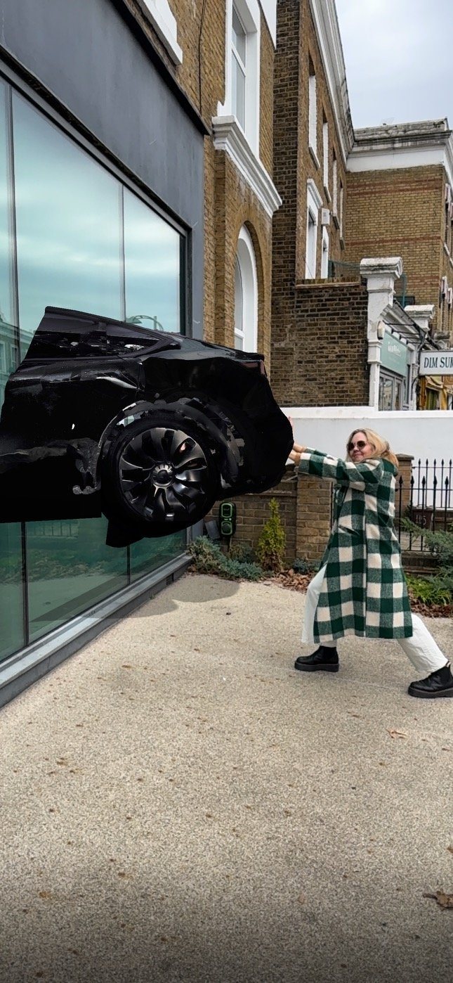 Woman in a green check coat pushes against a black car that appears to crash out of MOCA London's window, an AR view of Encounter.