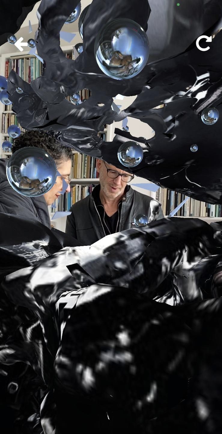 Encounter AR capture: two visitors peer at the black wreck from beneath a curl of twisted metal, chrome spheres beside them.